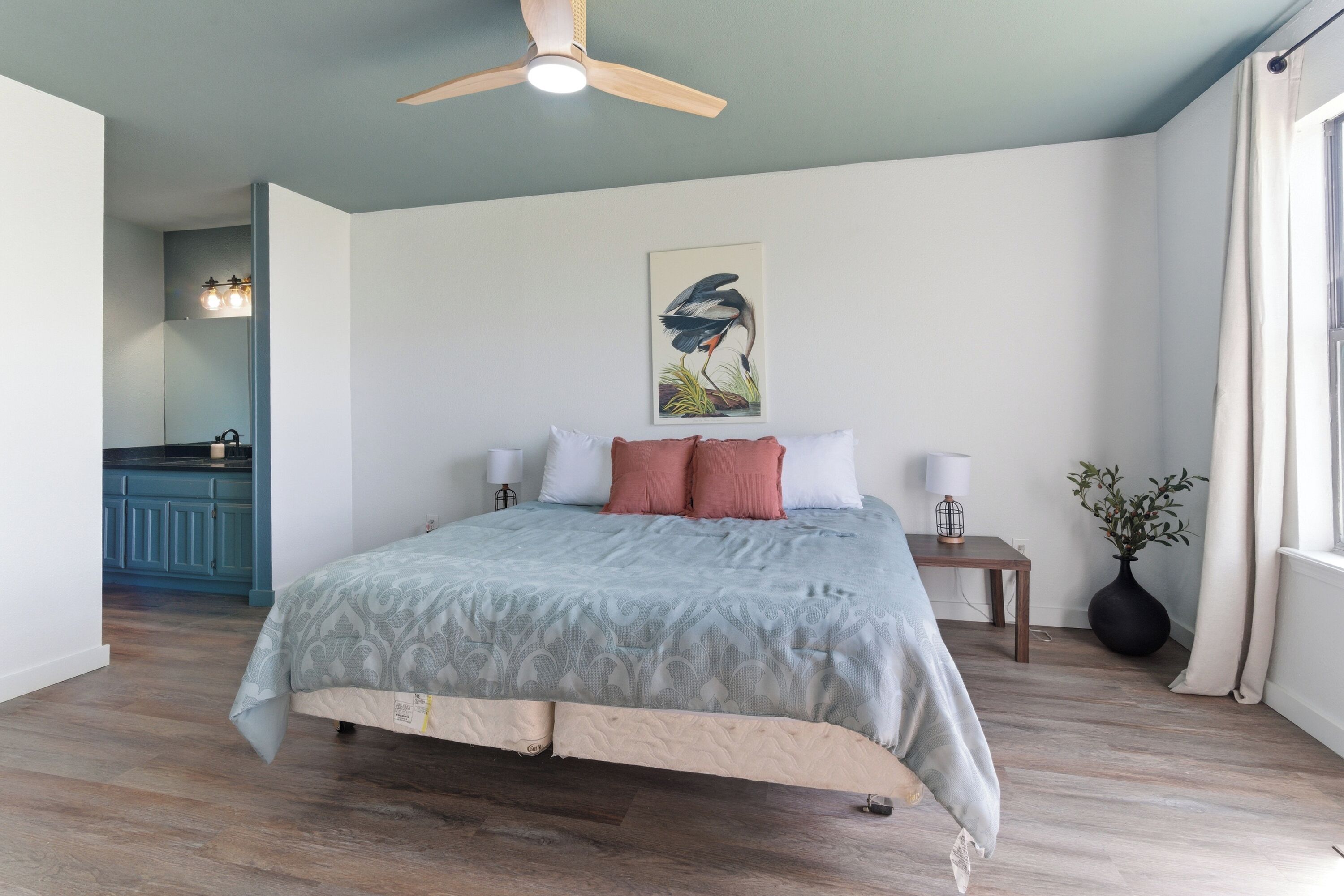 Une chambre à coucher comprenant un lit, un ventilateur de plafond, une table de chevet, une lampe, un tableau, une fenêtre et une plante.