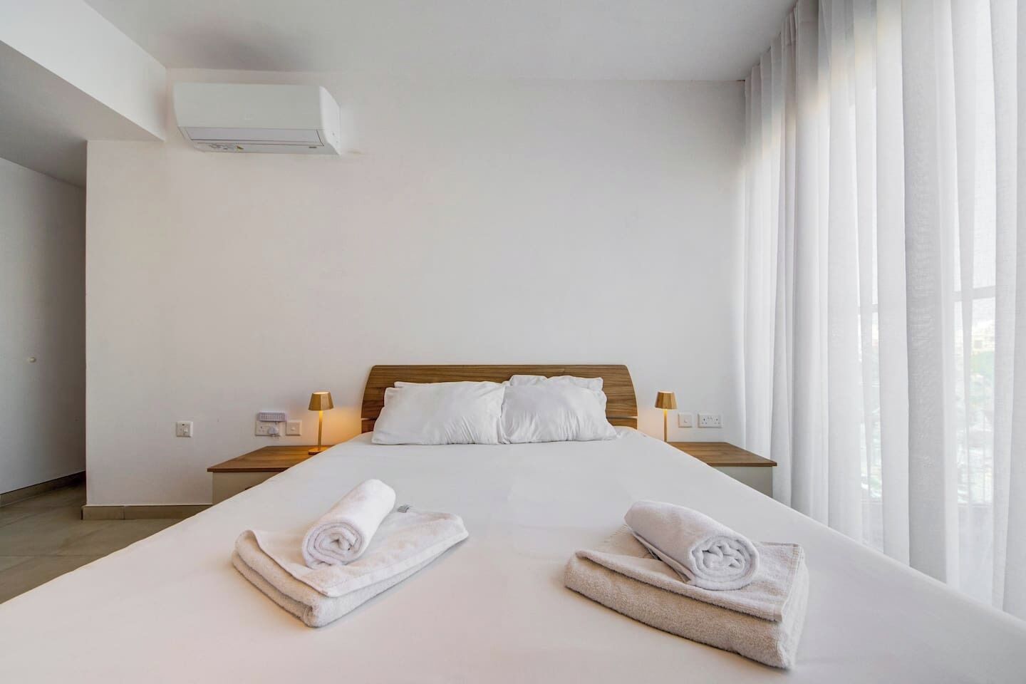 Une chambre d’hôtel avec un grand lit, deux tables de chevet avec des lampes, un climatiseur fixé au mur et une vue sur l’extérieur à travers des rideaux légers.
