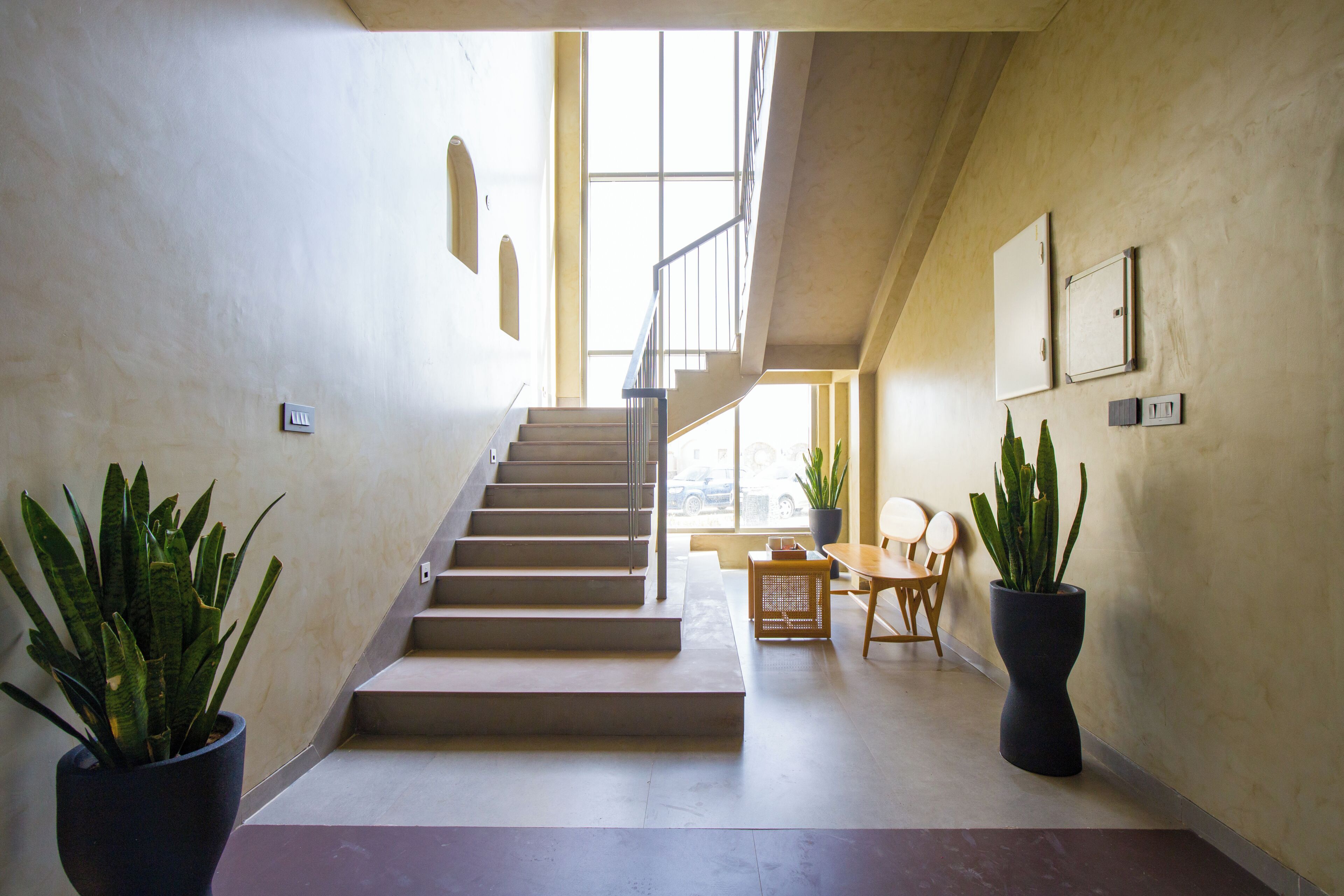 En trappe fører op til en etage, med plantekasser, et siddeområde med bord og stole, og et stort vindue der lukker naturligt lys ind.
