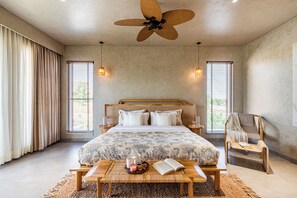 A bedroom with a large bed, a ceiling fan, two bedside lamps, a bedside table with a book and a tray of snacks, and a chair with a blanket.