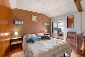 A bedroom with a bed, two bedside tables, a wooden wardrobe, a wicker chair, and a view through a glass door.