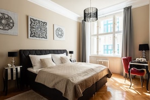 Une chambre à coucher avec un lit, des tables de chevet, un fauteuil rouge, une fenêtre avec des rideaux et des œuvres d’art murales.