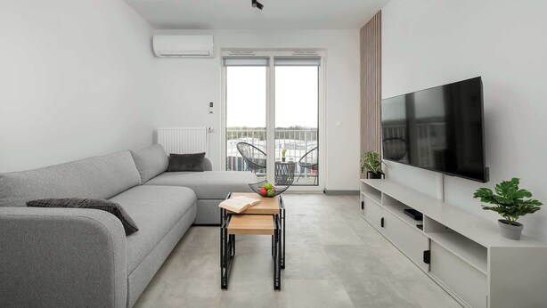 Una sala de estar moderna con un sofá, una mesita con un libro y fruta, un televisor y un balcón con sillas.