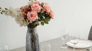 Una mesa de comedor dispuesta con platos blancos, copas de vino y cubiertos, junto a un jarrón con flores.