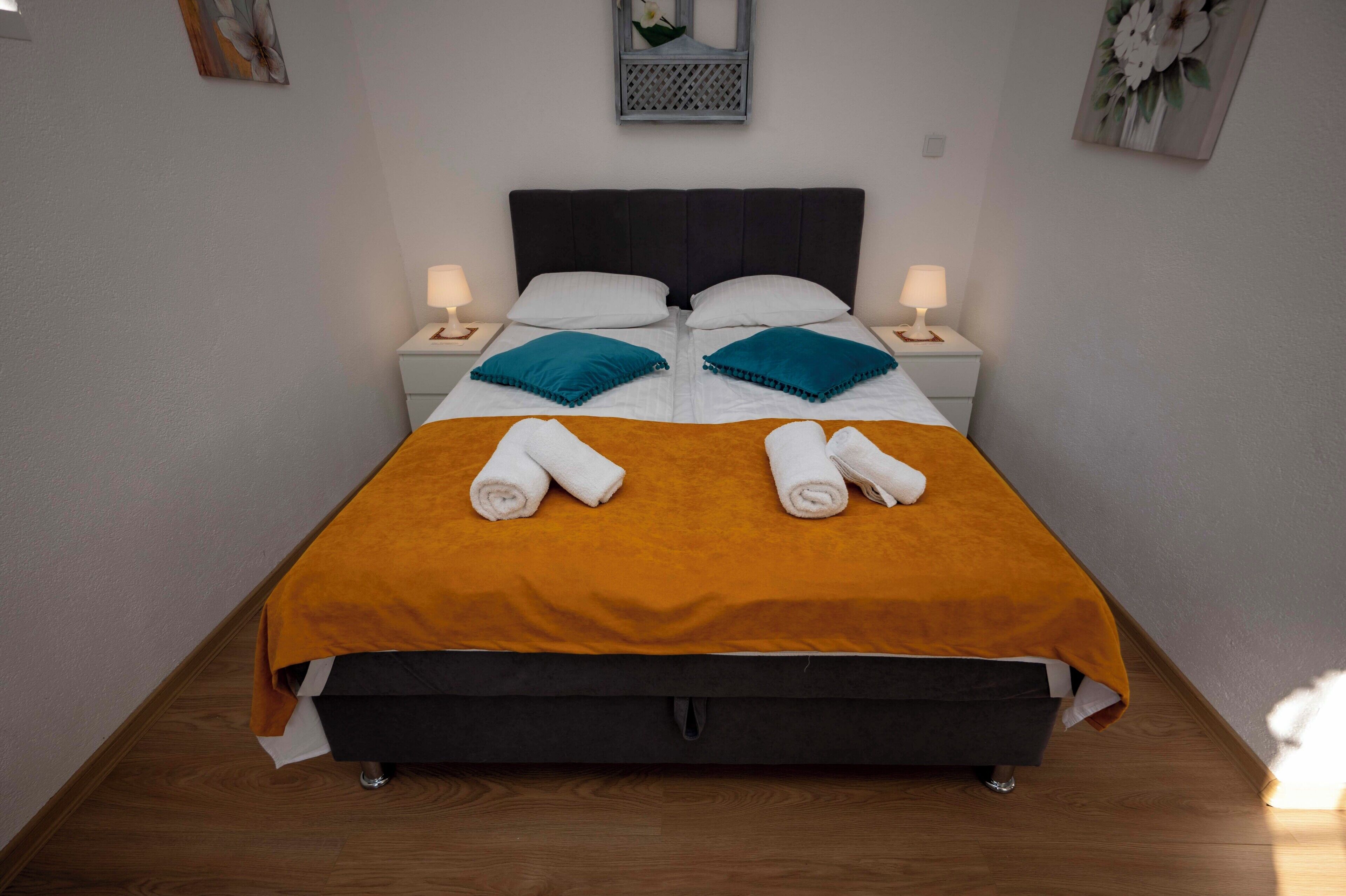 A neatly made bed with a dark headboard, two pillows, and two rolled towels.