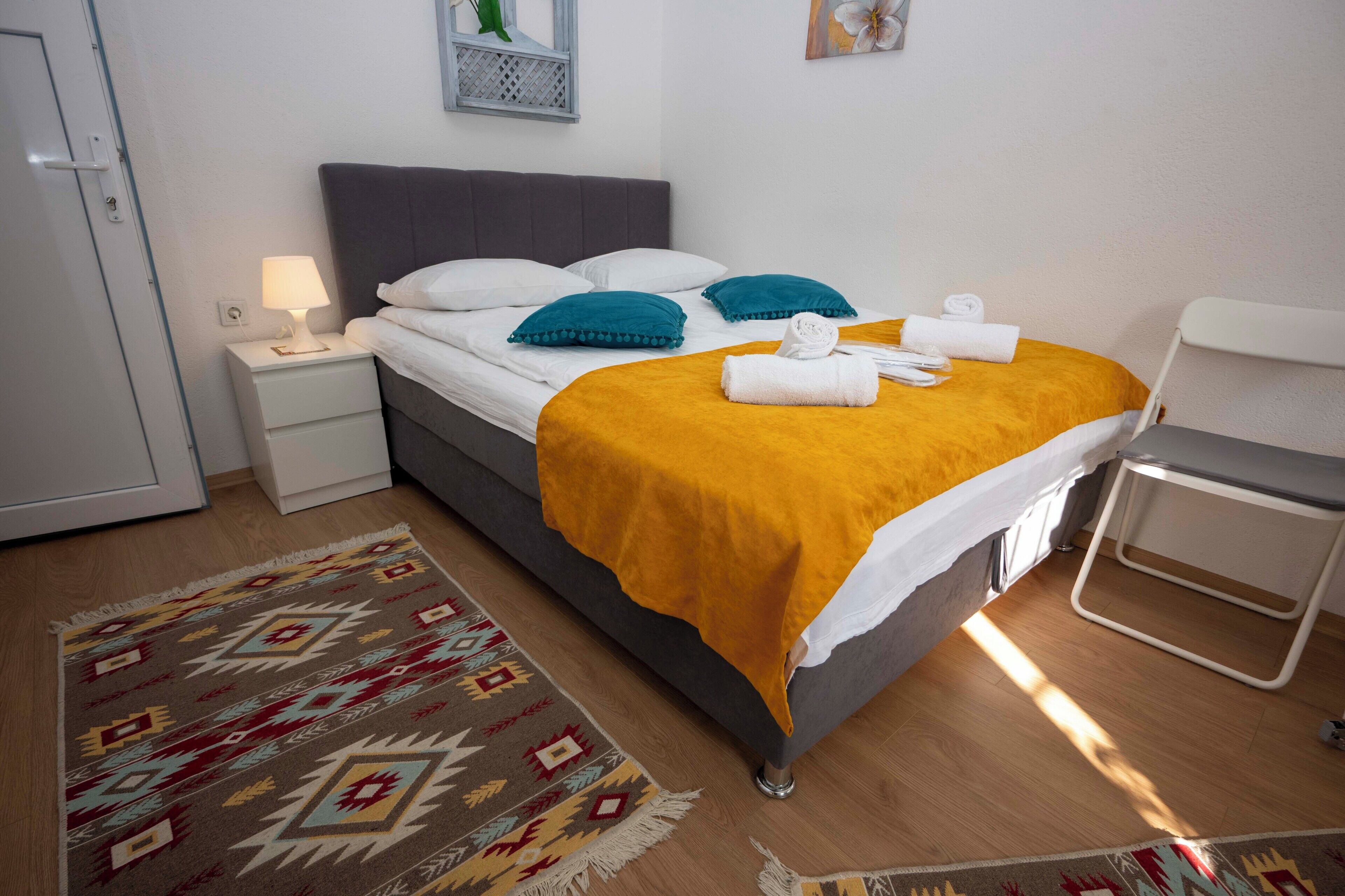 A neatly made bed with a yellow blanket, white pillows, and a blue accent pillow. There is a white bedside table with a lamp, a patterned rug, and a small white chair.
