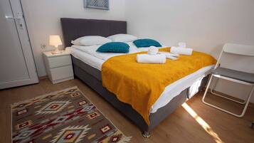 A neatly made bed with a yellow blanket, white pillows, and a blue accent pillow. There is a white bedside table with a lamp, a patterned rug, and a small white chair.