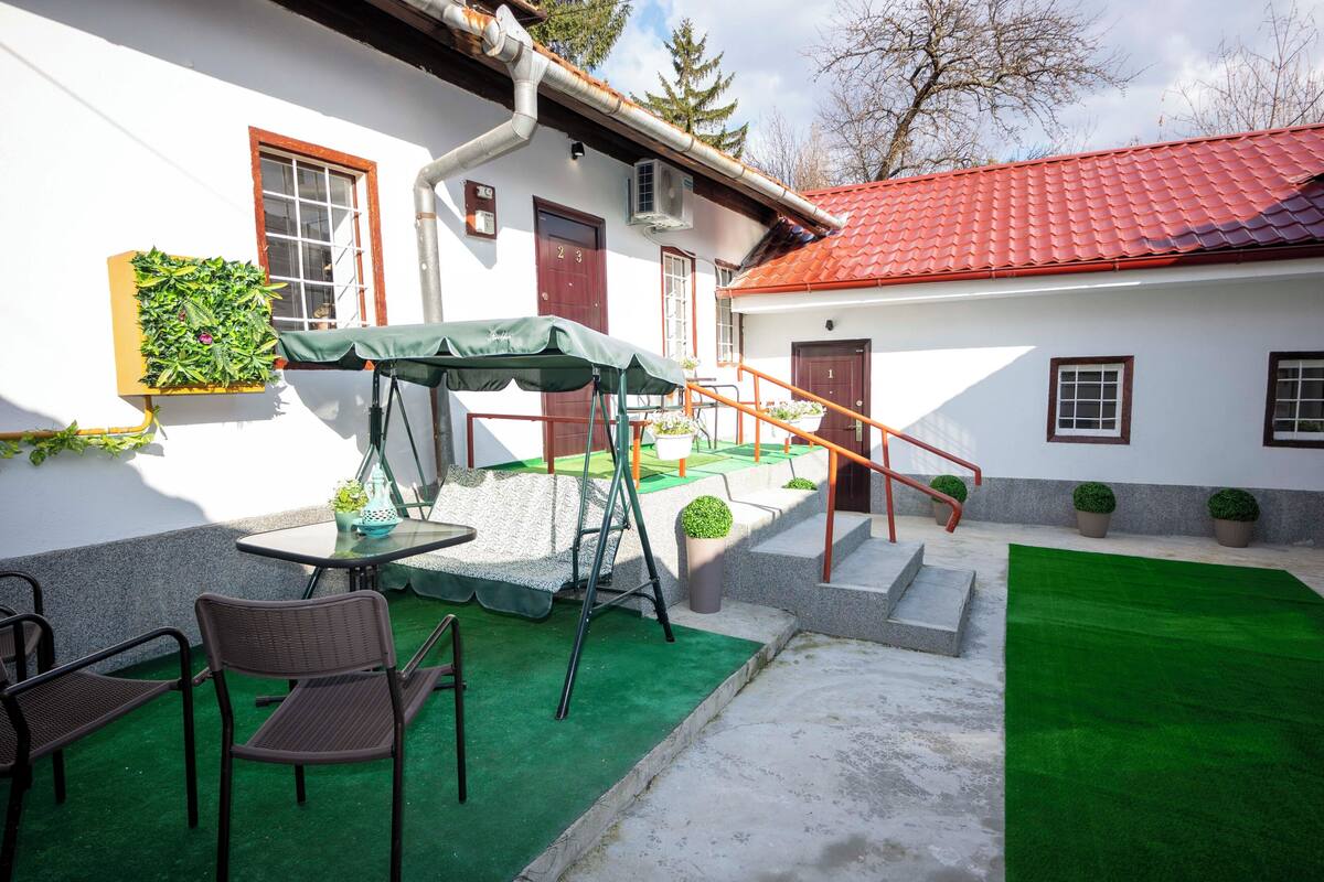 A white building with a red roof, a green outdoor seating area with chairs and a table, and a small staircase leading to a door.