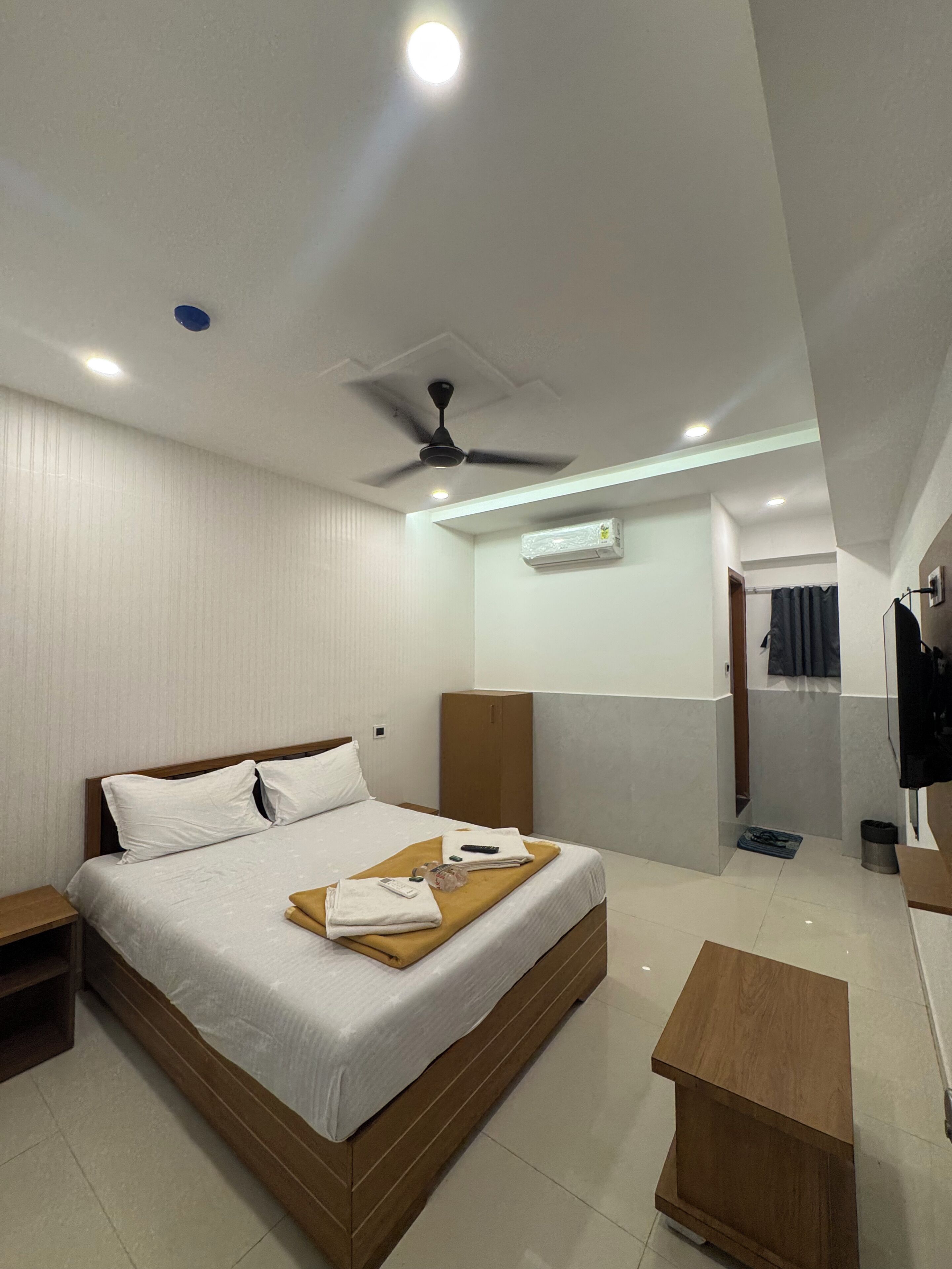 A hotel room with a bed, a ceiling fan, an air conditioning unit, a wooden bedside table, and a small wooden stool.