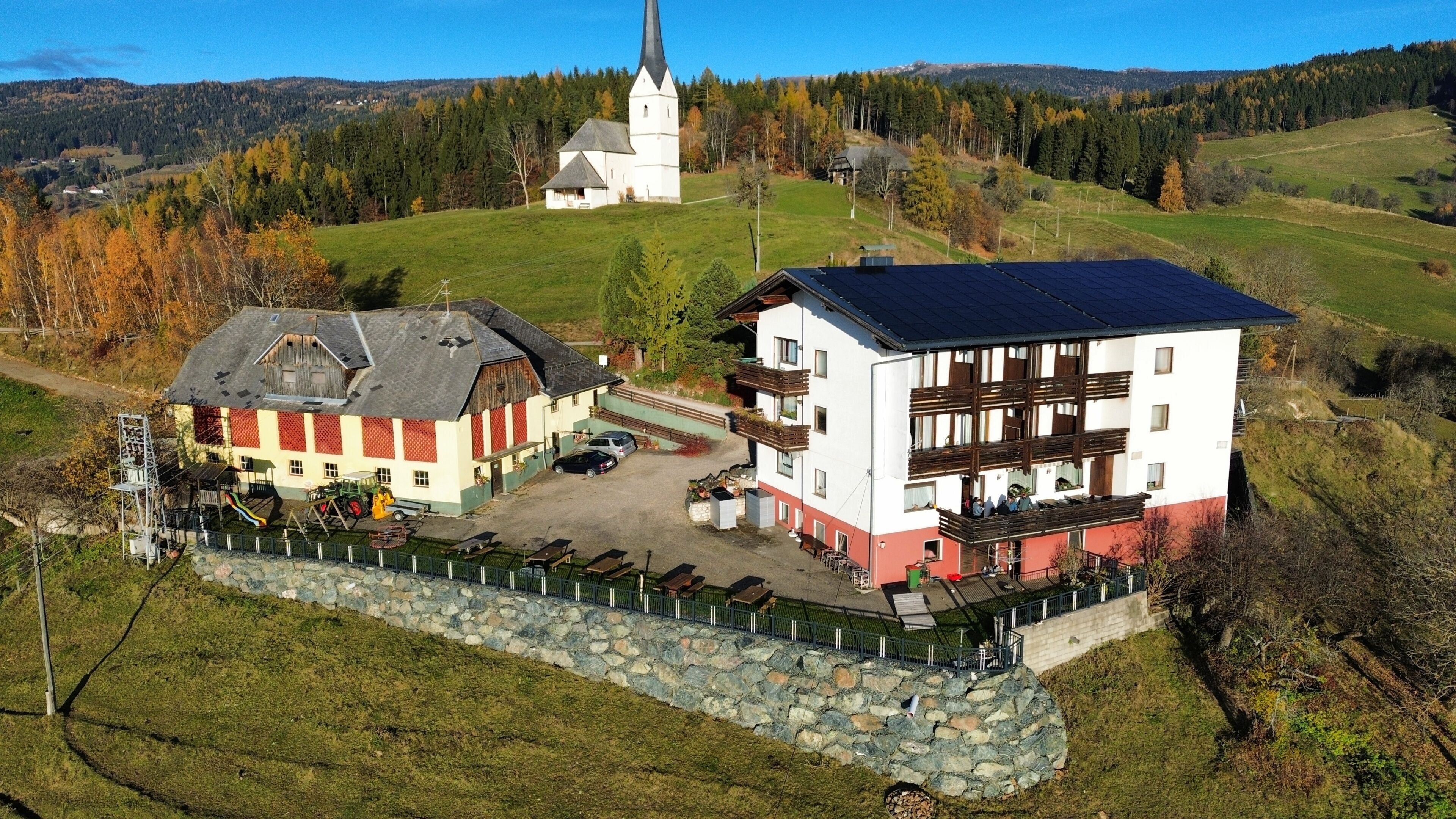 A multi-story building with a stone wall in front, a church in the background, and a green landscape.