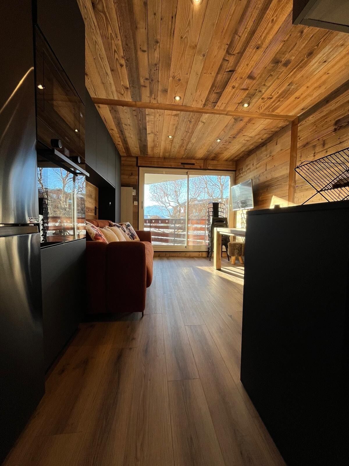 A modern kitchen with wooden ceiling beams, a large window, and a seating area with a sofa.