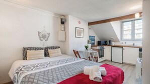 Une chambre avec un lit, une couverture rouge, un radiateur fixé au mur, une petite table avec une chaise et un coin cuisine.