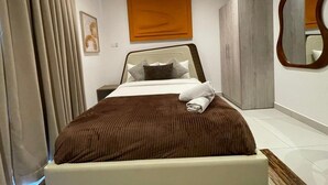 A bedroom with a bed, a nightstand, a mirror, and a wardrobe. - The Bantree by RoyalRest (Accra)