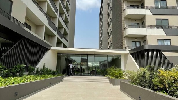 A modern building with multiple balconies, a central entrance with glass doors, and a landscaped area with plants. - The Bantree by RoyalRest (Accra)