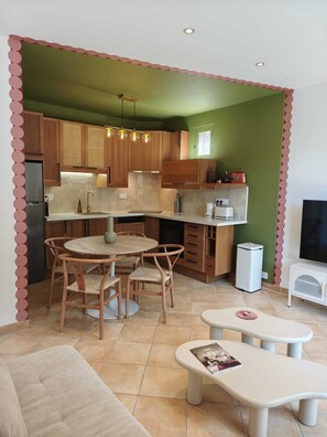 A modern kitchen with wooden cabinets, a dining area with a round table and chairs, and a small seating area with a bench and magazine. - Horizon Apartment (Ota)