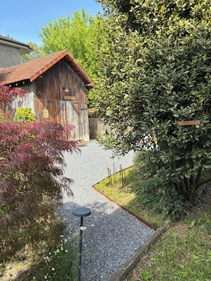 A rustic wooden barn with a gravel driveway leading to it, surrounded by dense shrubbery and a variety of plants.