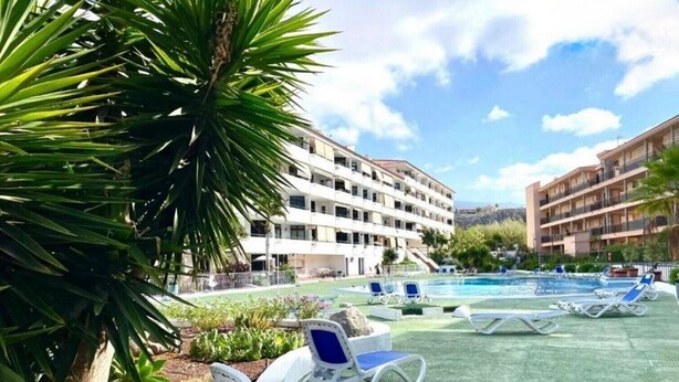 A pool area with lounge chairs, a building with multiple balconies, and a palm tree. - Summerland 5 - 1 bedroom with terrace and pool view (Los Cristianos)