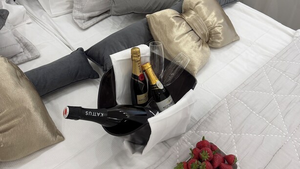 Ein ordentlich bezogenes Bett mit grauem Kopfteil, weißen und grauen Kissen, einem schwarzen Korb mit Champagnerflaschen und einer Schale Erdbeeren.