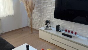 Ein modernes Wohnzimmer mit einem Flachbildfernseher, einer weißen Kommode mit roten Kerzen, einer Obstschale und einer Weinflasche.