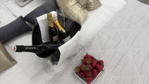 Ein ordentlich bezogenes Bett mit weißer Bettdecke, grau-goldenen Kissen, einer Champagnerflasche und einer Schale Erdbeeren.