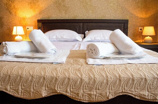 A hotel room with two beds, each with a rolled white towel and a folded white bath mat, a dark wooden headboard, and two bedside lamps.