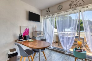 Une pièce avec une table en bois, des chaises, un téléviseur fixé au mur, une fenêtre avec des rideaux légers et une petite table sur laquelle se trouvent une lampe et un livre.