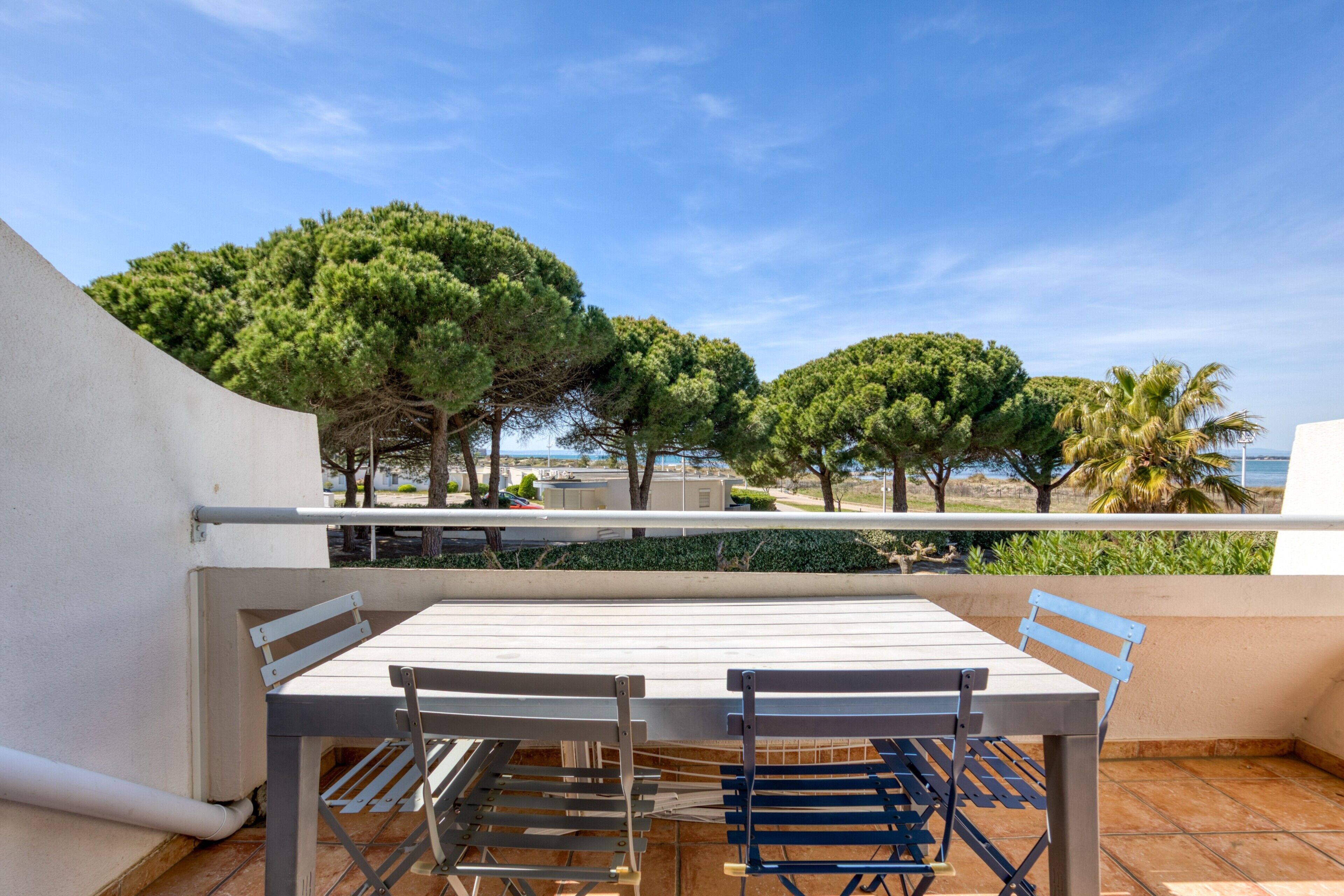 A balcony with a table and chairs, overlooking a coastal area with trees and a clear sky.