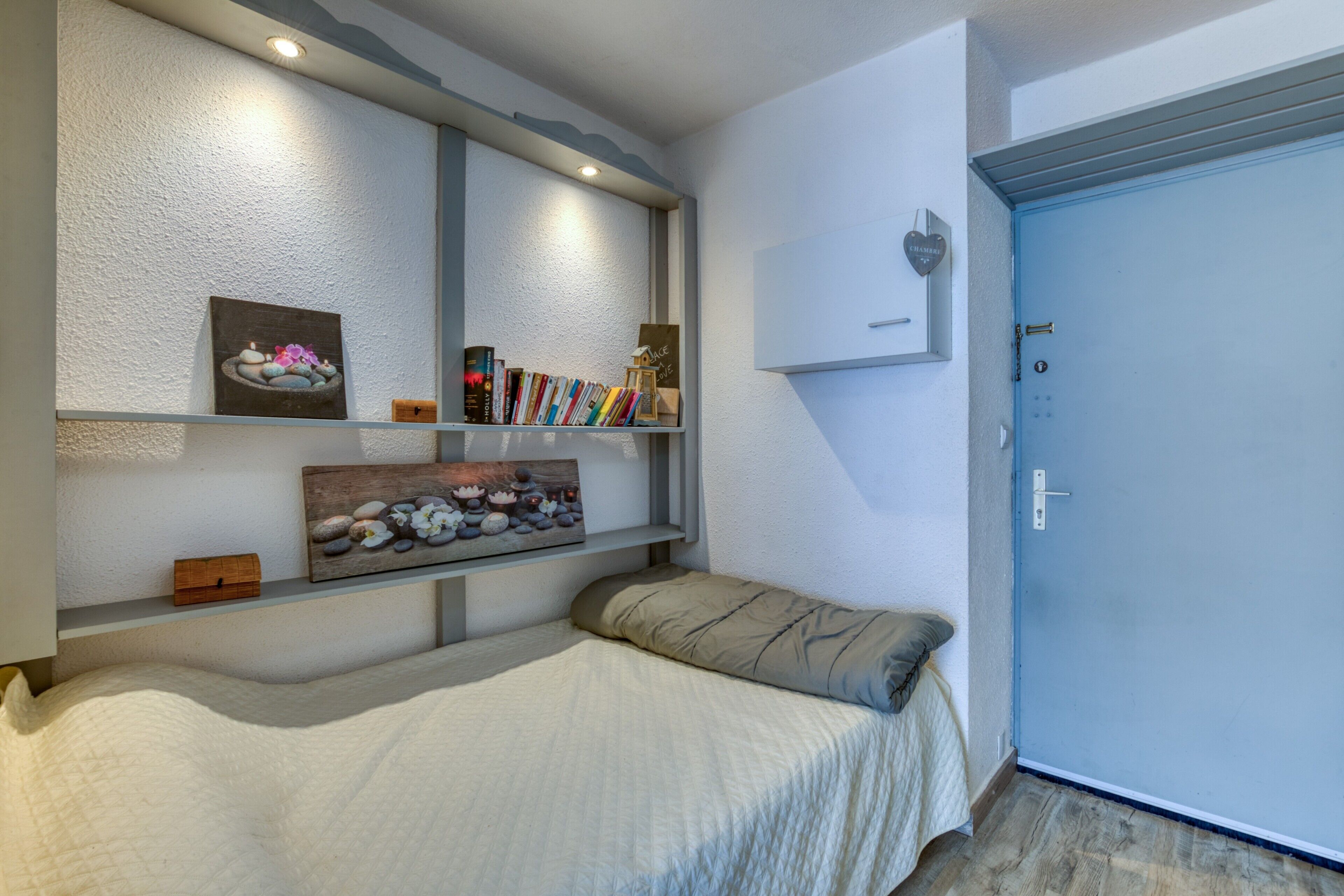 A bedroom with a bed, a bedside table, a shelf with books and decorative items, a door, and a wall-mounted light fixture.