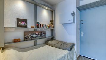 A bedroom with a bed, a bedside table, a shelf with books and decorative items, a door, and a wall-mounted light fixture.