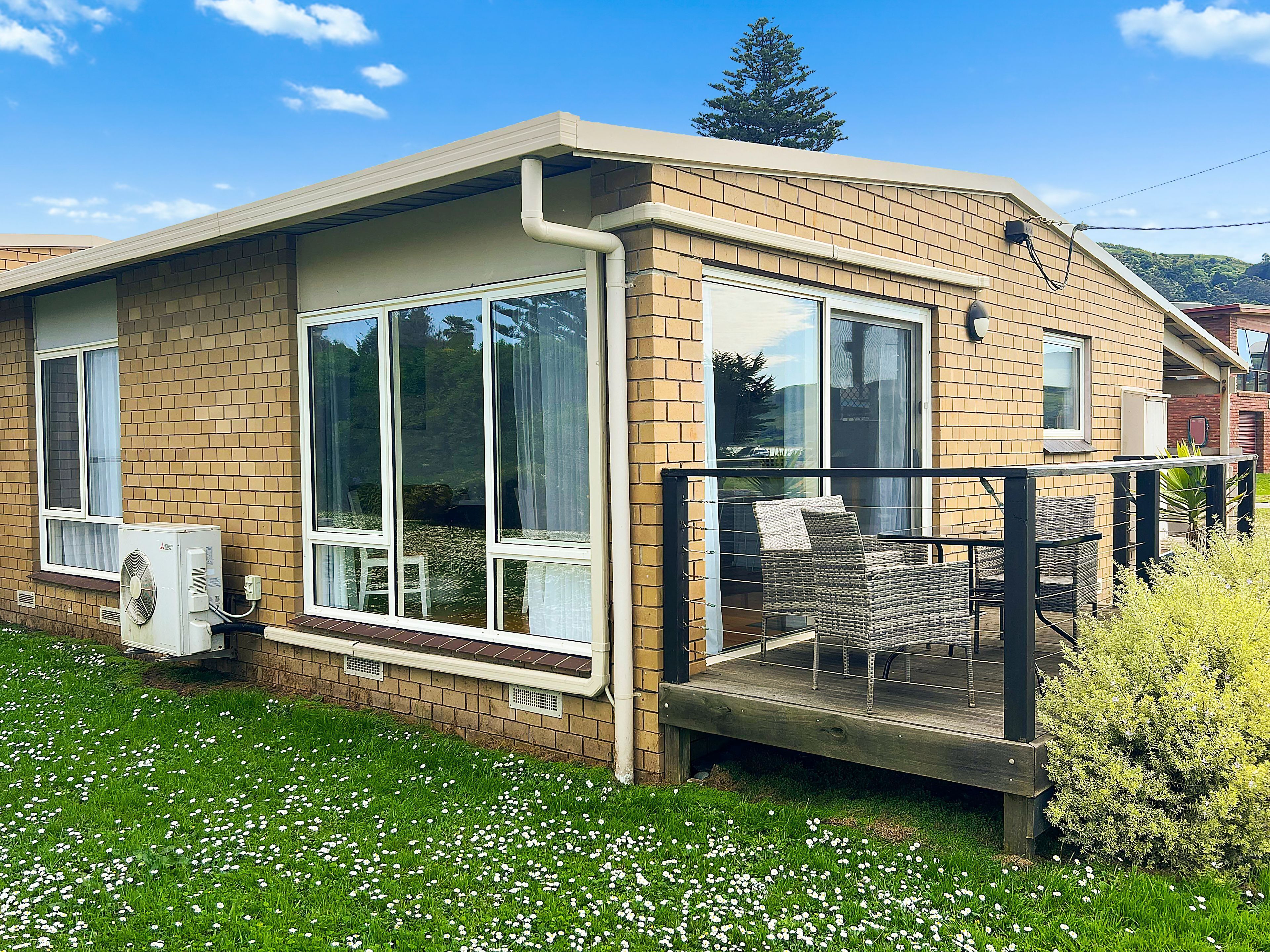Une maison de plain-pied avec une façade en briques, de grandes fenêtres et une véranda couverte aménagée avec du mobilier d’extérieur.