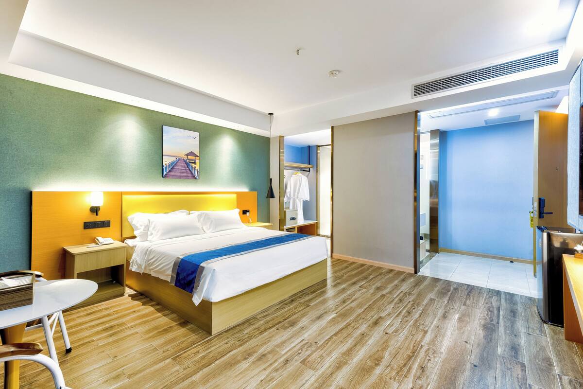 Een hotelkamer met een bed, nachtkastje, lamp en uitzicht op de gang.
