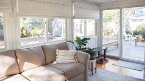Ett rum med en beige soffa, stora fönster med persienner, en krukväxt och utsikt över en terrass och hus utanför.