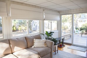 Ein Zimmer mit einem beigen Sofa, großen Fenstern mit Jalousien, einem Blumentopf und Blick auf eine Terrasse und Häuser draußen.