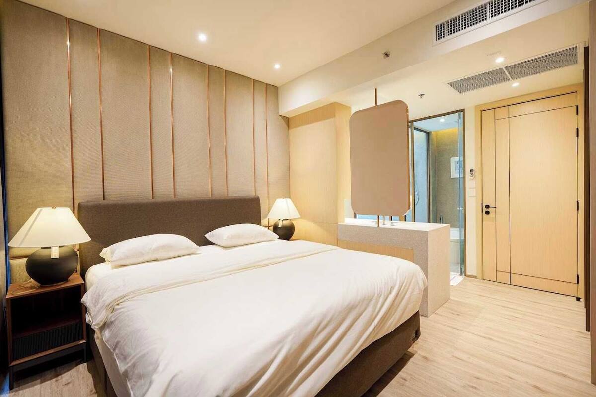 A hotel room with a large bed, two bedside tables with lamps, a wardrobe, and a bathroom visible through a glass door.
