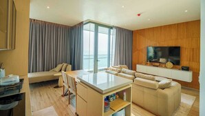 A modern living room with a sofa set, a dining area, a TV mounted on a wooden wall, and a view of the sea through large windows. - Luxury 2BR Sea View Apartment with Private Lift and Free Parking (Pattaya)