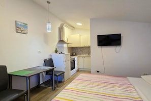 A compact room with a bed, a desk with a chair, a kitchenette, and a TV mounted on the wall. - Studio Apartment Nia (Zaton)