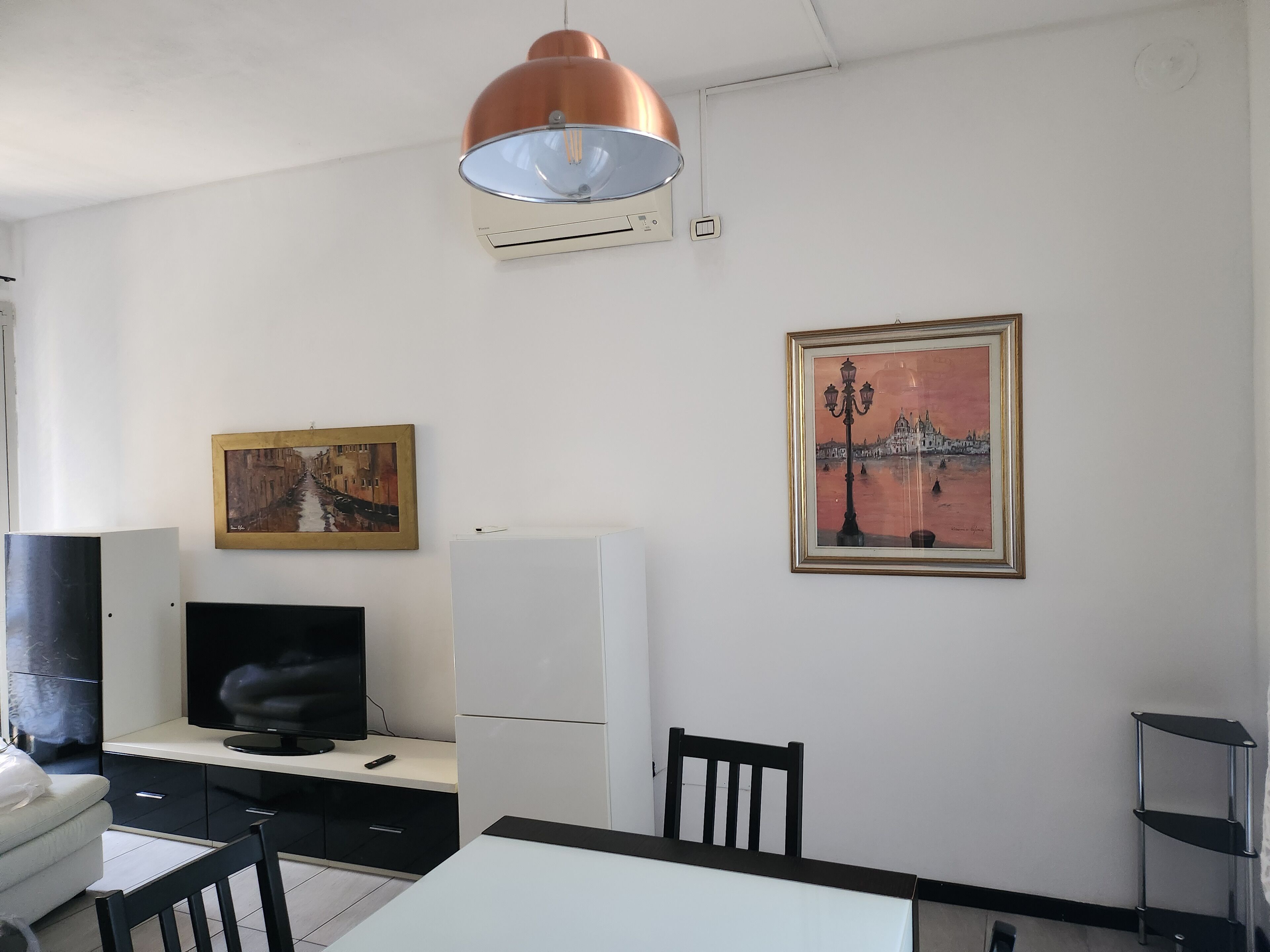 Een moderne woonkamer met een flatscreen-tv, een eettafel, stoelen, een witte bank en twee ingelijste schilderijen aan de muur.