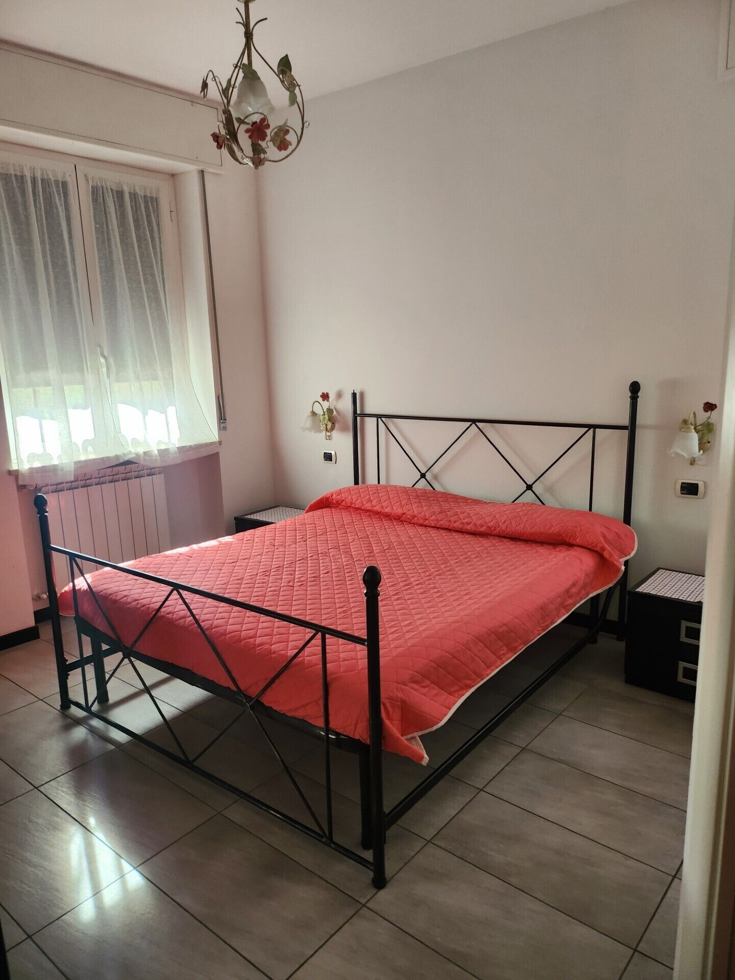 Una camera da letto con una struttura letto in metallo, una coperta rossa, una finestra con tende trasparenti, una lampada a parete e un comodino.