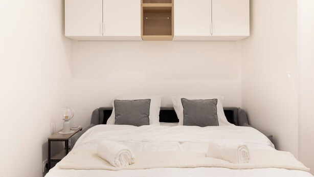 A neatly made bed with white bedding, a bedside table with a lamp, and a wardrobe above the bed. - Suite Élégance at Montparnasse (Paris)