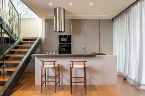 Eine moderne Küche mit Kochinsel, Barhocker, einem Edelstahl-Dunstabzug und einer Treppe mit Glasgeländern.