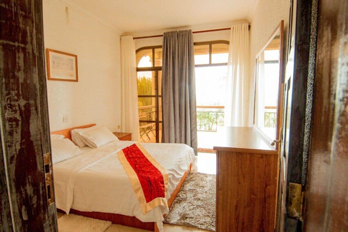 Une chambre à coucher avec un lit, une table de chevet en bois, un bureau, une fenêtre avec des rideaux et une vue sur l’extérieur.