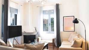 A living room with a flat-screen TV, a fireplace, a sofa, a coffee table, and a window with curtains. - Hebe's house | Cozy Village Escape Near Tripoli (Stadio)