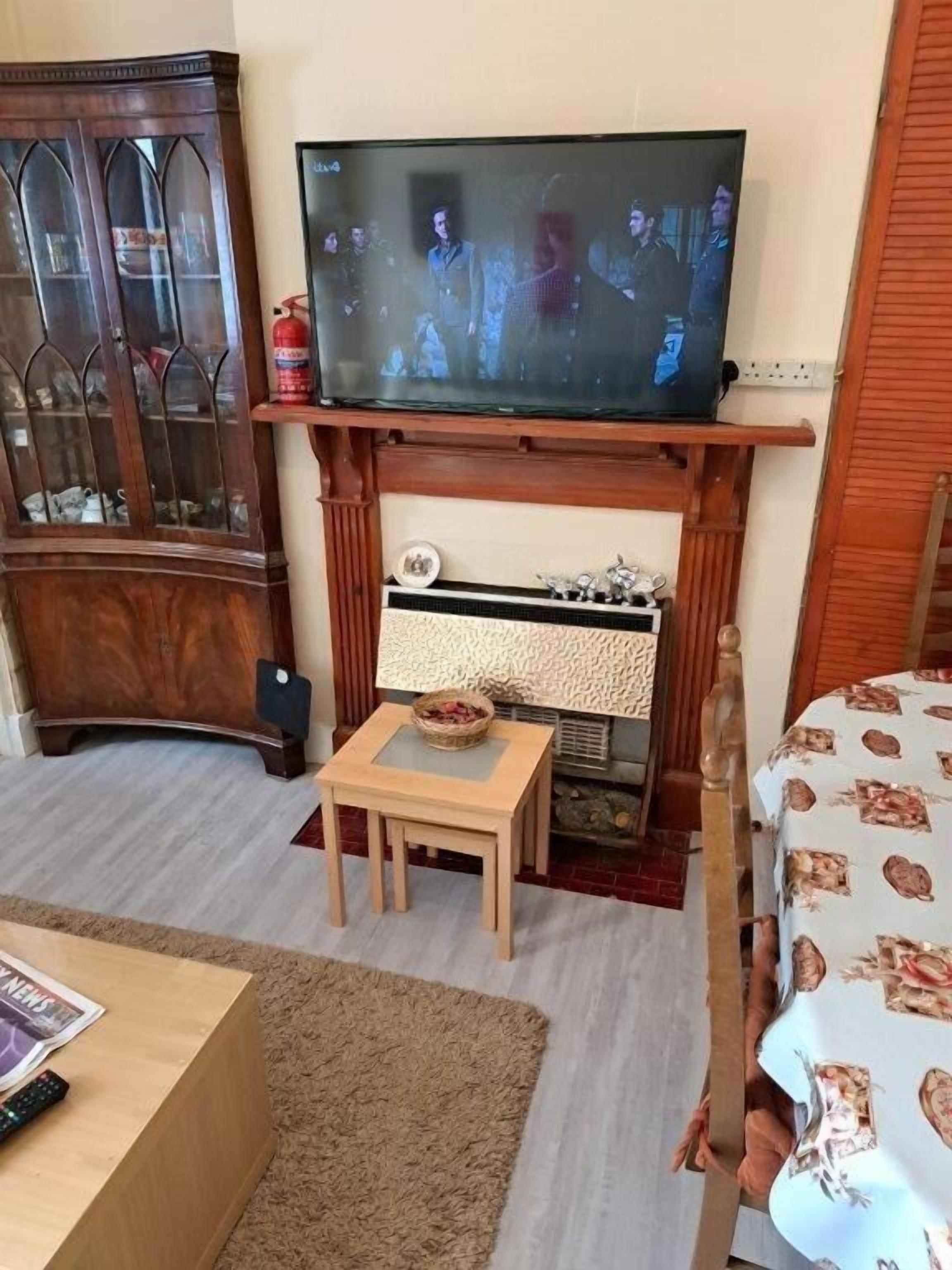 A living room with a television on a stand, a wooden cabinet, a fireplace, a small table, and a bed with a floral bedspread.