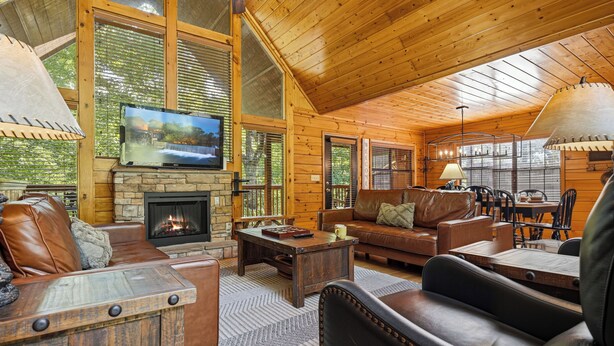 A log cabin interior with a stone fireplace, leather furniture, a television, and large windows. - New Listing, Game Room. Hot Tub, Pool Access +Free Attractions! (Sevierville)