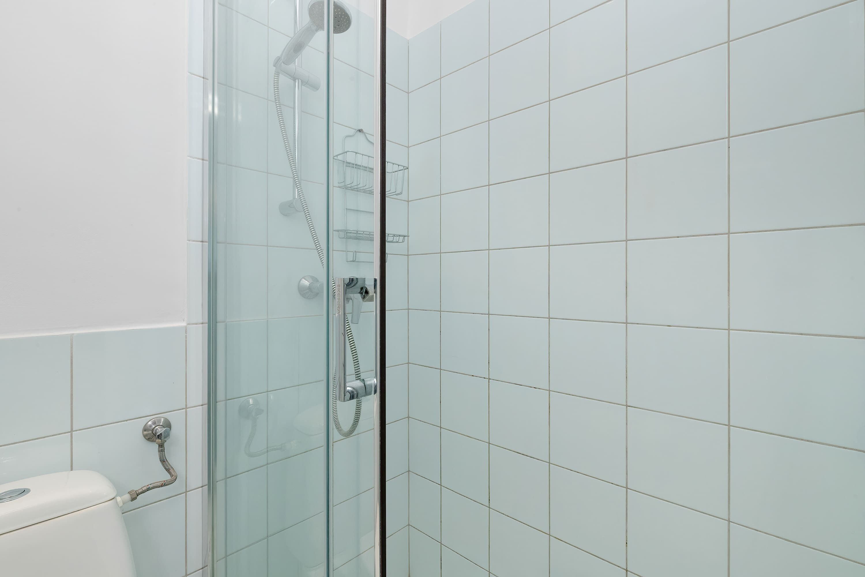Ducha con mampara de vidrio, cabezal y manguera de cromo, plato de ducha de vidrio y pared con azulejos blancos.