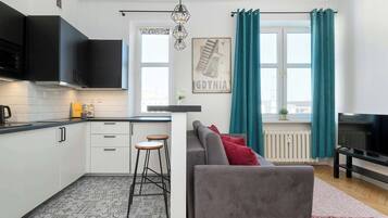 Moderni keittiö, jossa on mustat kaapit, valkoinen työtaso ja sisäänrakennettu mikroaaltouuni. Olohuoneessa on harmaa sohva, jossa on punaiset koristetyynyt, televisio ja ikkuna, jossa on turkoosinsiniset verhot.
