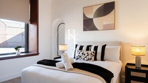 A bedroom with a bed, bedside tables, a lamp, a window with a view, and a wall-mounted abstract painting. - The Den on Hay Crescent (Peterhead)
