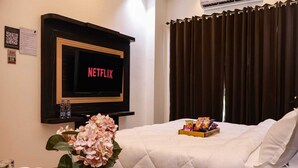 A hotel room with a bed, a TV displaying the Netflix logo, a bouquet of flowers, snacks, and a water bottle. - The Glam By Sandane Homes (Greater Noida)