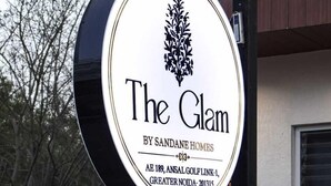 A sign for 'The Glam' by Sandane Homes, featuring a decorative emblem and address details. - The Glam By Sandane Homes (Greater Noida)
