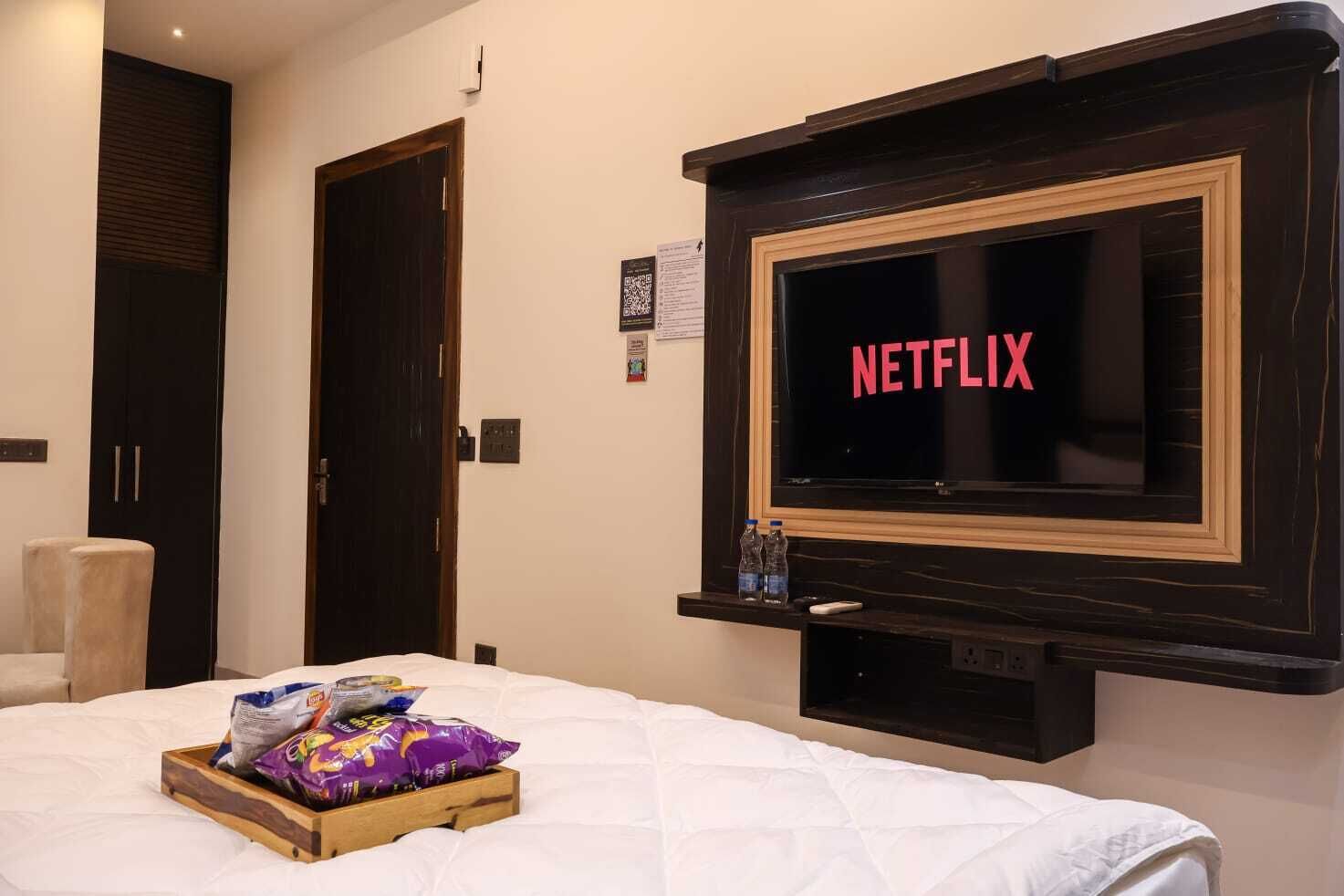 Quarto de hotel com cama, TV na parede exibindo 'NETFLIX', bandeja de madeira com lanches e uma cadeira branca.